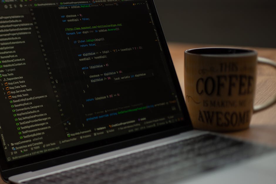 A close-up view of a laptop on a wooden surface displaying lines of code in a text editor, with a beige ceramic mug positioned behind the laptop. The mug has text that reads ' THIS COFFEE IS MAKING ME