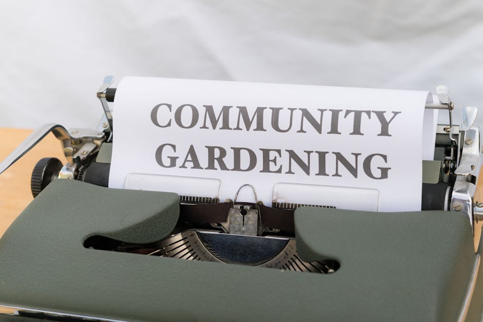 A vintage green manual typewriter placed on a wooden surface, with a sheet of paper inserted that displays the words 'COMMUNITY GARDENING' in bold, uppercase letters. The background is neutral and out